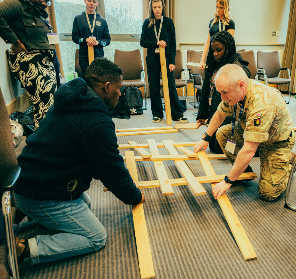 Participants take part in a team-building workshop, arranging wooden planks on the floor as part of a practical challenge. One participant in military uniform kneels alongside another participant to position the pieces, while several others stand and watch around the room. Chairs are arranged in a circle, and the activity takes place in a bright meeting room with large windows.