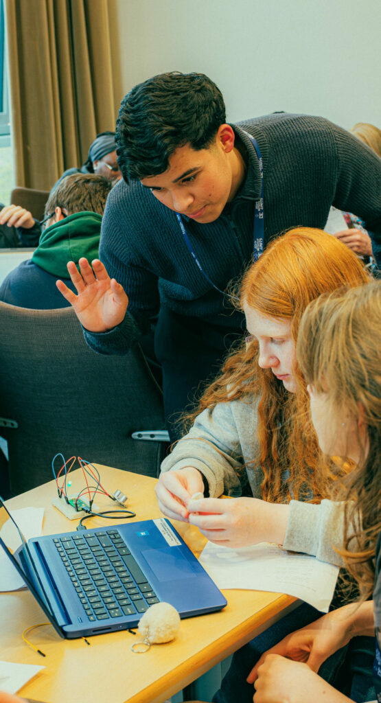 Participants work together at a table during a hands-on technology workshop. A laptop, electronic components, wires and printed instructions are spread across the table as one participant leans over to assist others with the activity. Additional groups can be seen collaborating in the background of the workshop room.