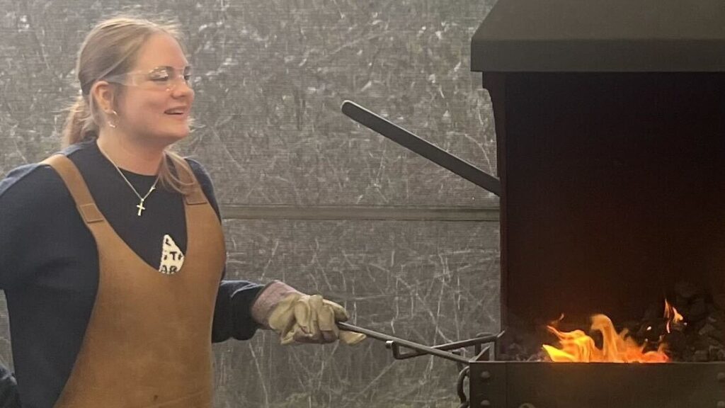 Amy is standing by an open fire wearing a brown apron.