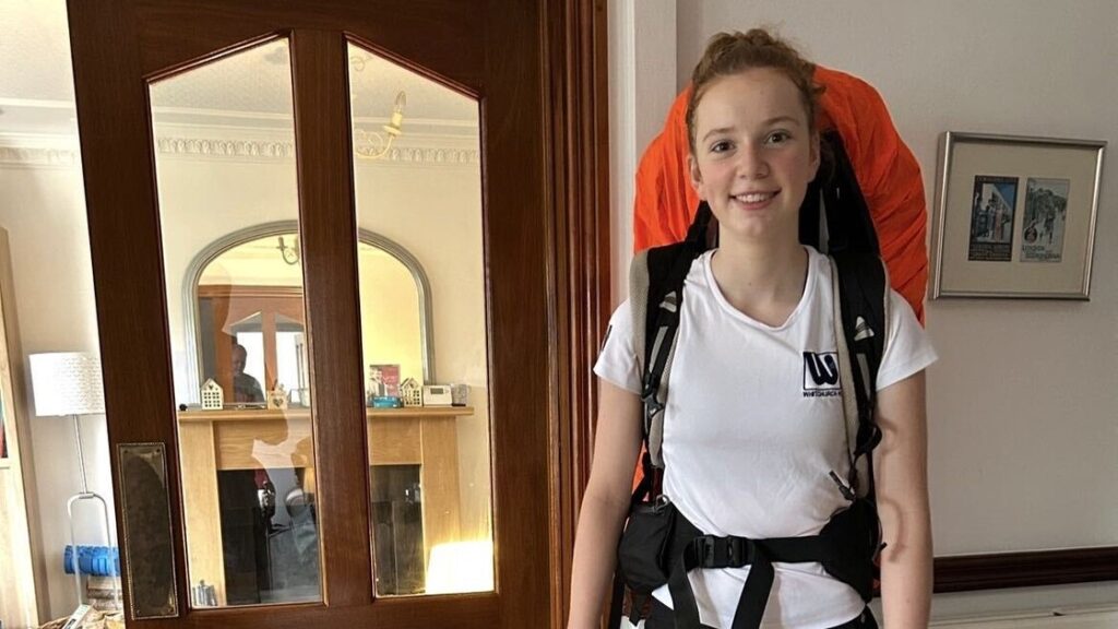 Anna is smiling and wearing a big rucksack indoors.