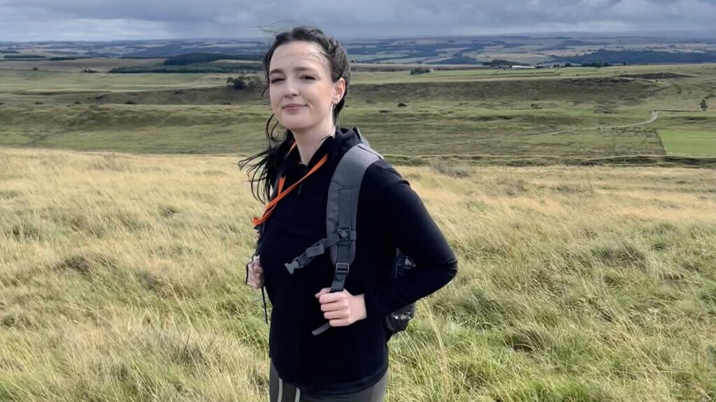 Caitlin is standing on a grassy field in front of a landscape view.