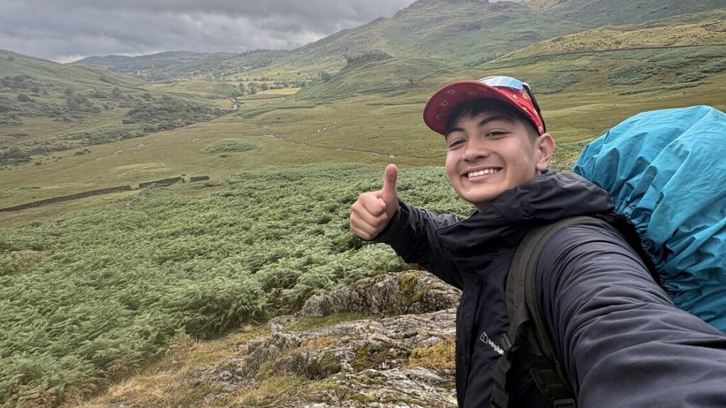 Dan is taking a selfie smiling with a thumbs up carrying a rucksack outdoors.