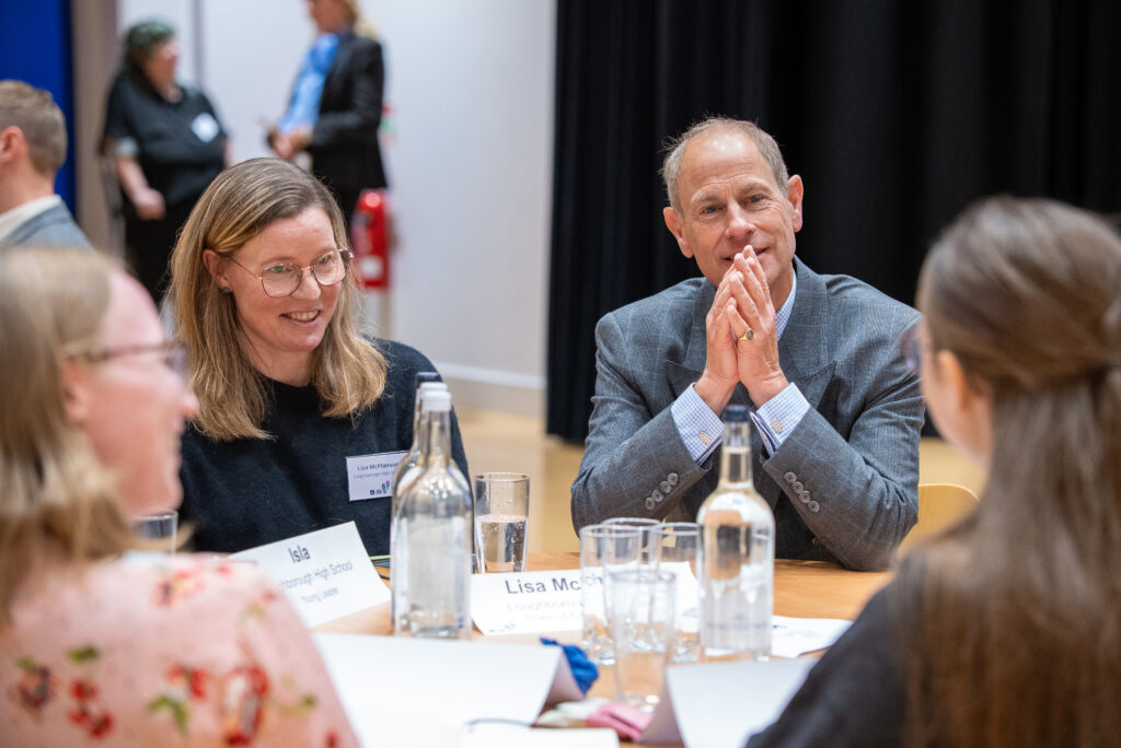 HRH The Duke of Edinburgh sits at a roundtable discussion, listening to participants seated around a table with water bottles and name cards.