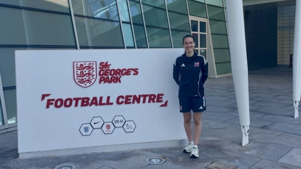 Ella is wearing black and white sports kit standing next to a sign that says "St George's Park football centre".