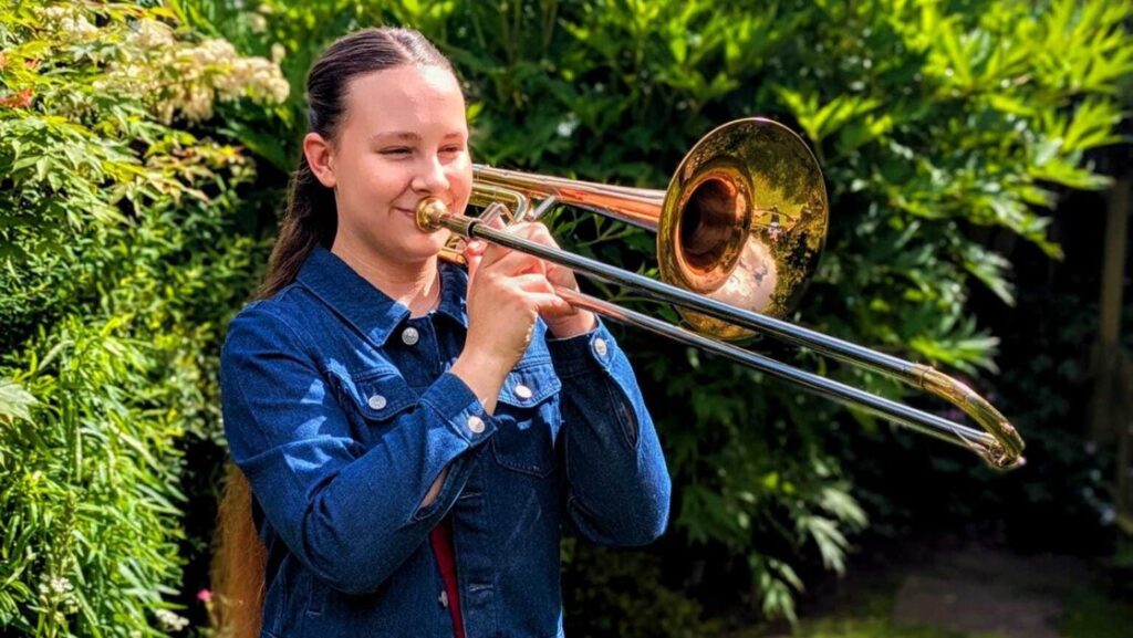 Evelyin is standing in the garden playing the trombone.