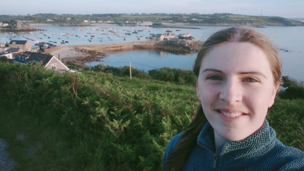 Evie has dark blonde hair in a side ponytail and a blue jacket, smiling in front of a coastal landscape.