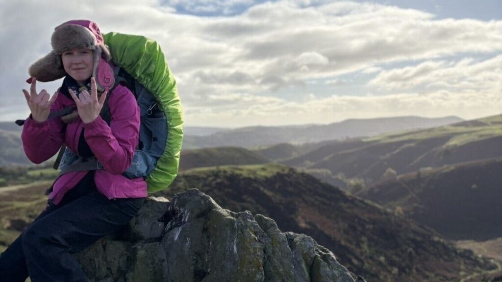 Fifi sits on a rock atop a mountain wearing a pink hat and jacket, carrying a big, green rucksack.