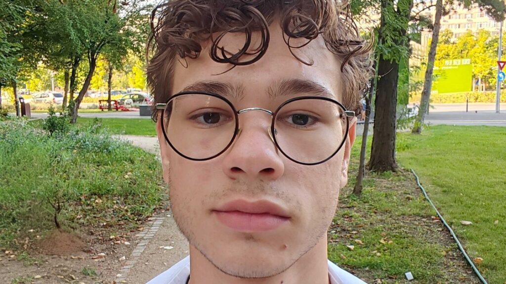 Laurentiu has round, black glasses and brown curly hair, standing in a park.