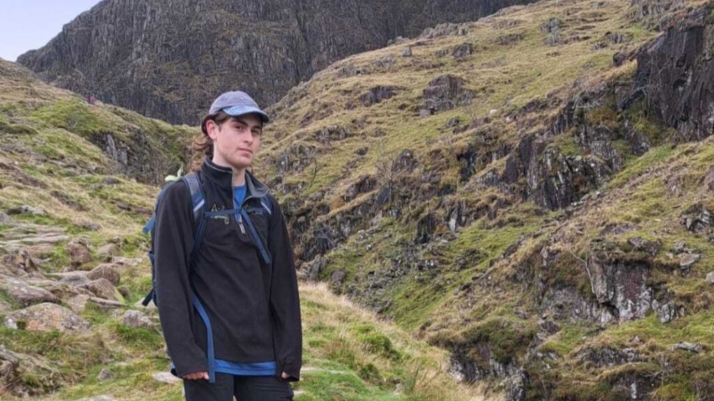 Leo is wearing a hat and black jacket, standing on a mountain path.