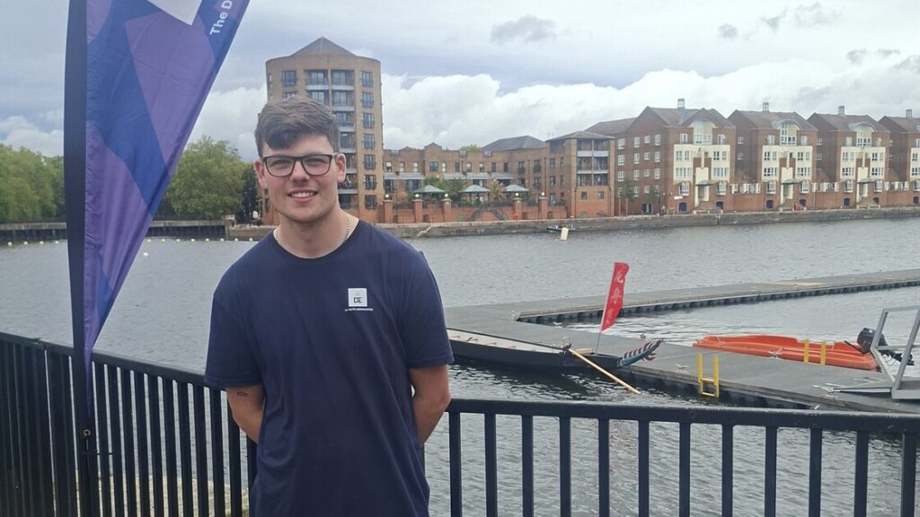 Lewis is smiling, standing in front of a river with a dragon boat on it.