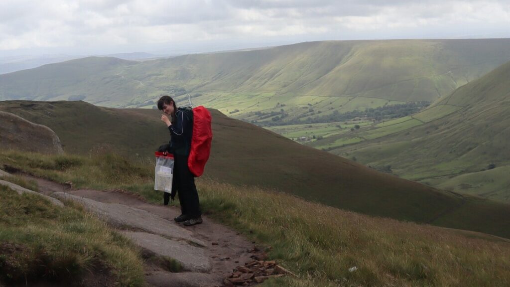 Lucy is carrying a big, red rucksack, standing on a mountain path in front of a view of hills.