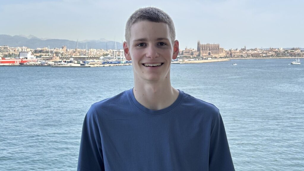 Luke is smiling and wearing a blue t-shirt in front of an ocean view.