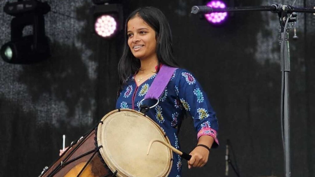 Parisha wears a blue patterned dress playing a large drum on a stage with lights in the background.
