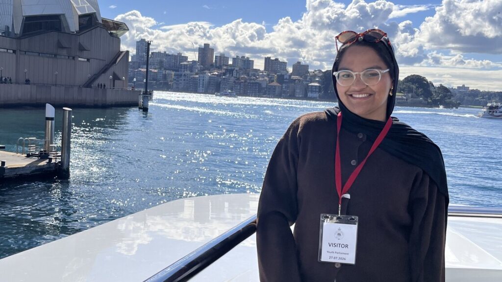 Qahira is smiling, standing in front of a city skyline on the water.