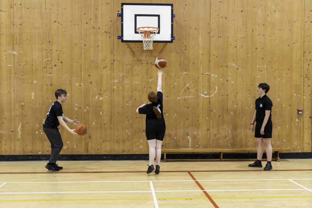 A basketball practice scene in a gymnasium with a wooden wall. A girl with red hair is shooting a basketball towards the hoop, while a boy in glasses stands nearby, holding a basketball and preparing to pass. Another boy watches from the side, both wearing black athletic shirts. The gym floor is marked with lines for basketball.