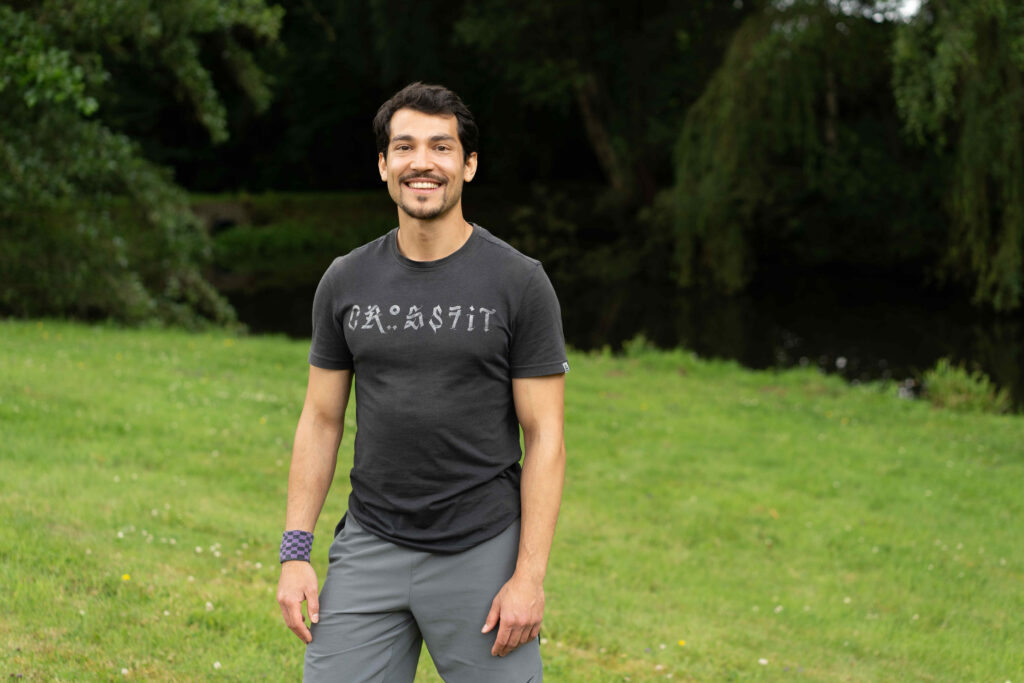 Lionel Bush stands outdoors on a grassy area near a body of water. He is wearing a dark gray t-shirt with a graphic design and light gray shorts. The background features trees and greenery, creating a natural setting.