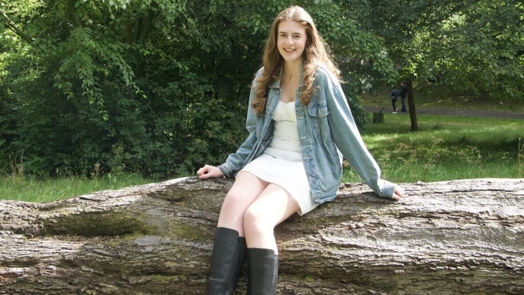 Sarah is wearing a white dress and denim jacket sitting on a big log in a garden.