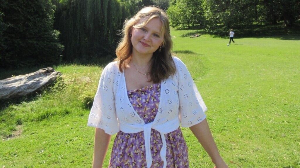 Tabitha is wearing a white jacket and floral dress standing in a garden smiling.