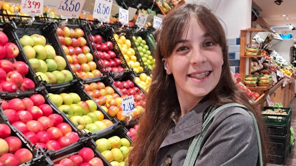 Tegwen has brown hair with a fringe, she's standing in a supermarket smiling.