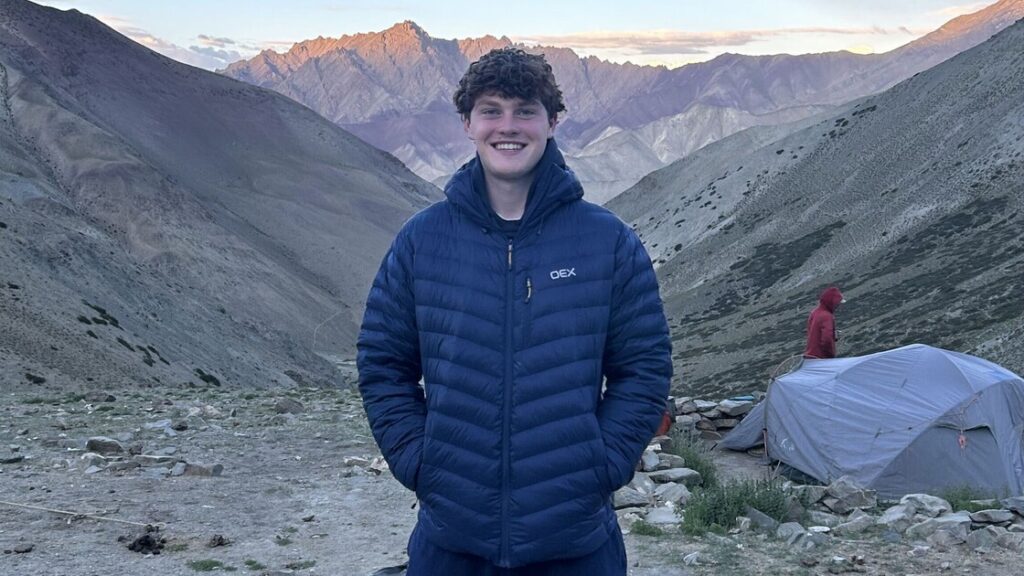 Tristan is standing in the mountains wearing a blue puffer jacket, there's a grey tent behind him.