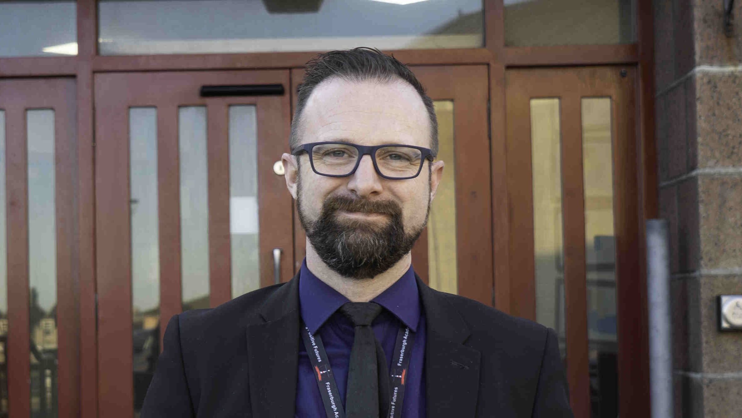 A man in a suit stands in front of Fraserburgh Academy, which has a sign that reads "Welcome to Fraserburgh Academy" above the entrance. The building features a modern design with large windows and a welcoming entrance. The man is smiling and appears confident, with a lanyard around his neck.