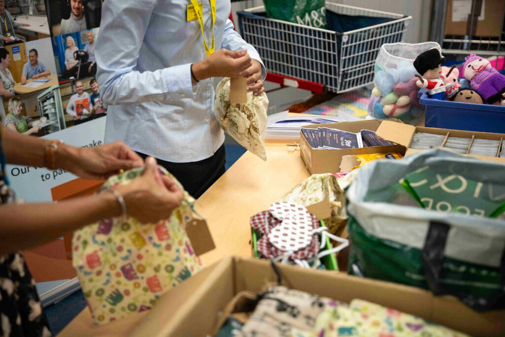 A young woman in a white shirt hands a stack of colorful knitted items to a nurse in a blue uniform at a reception desk in a hospital setting. The background features computers and a welcoming environment, indicating a healthcare facility.