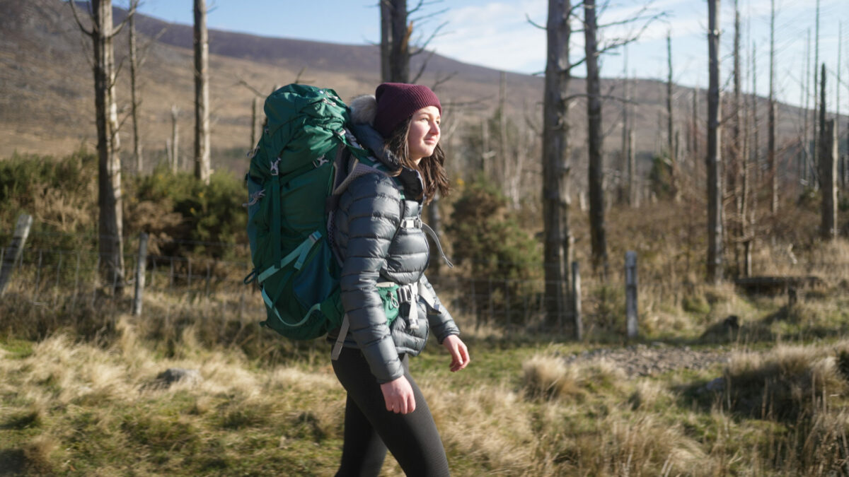 A young person carrying a rucksack, wearing a puffy jacket and red hat walks on a mountain path.