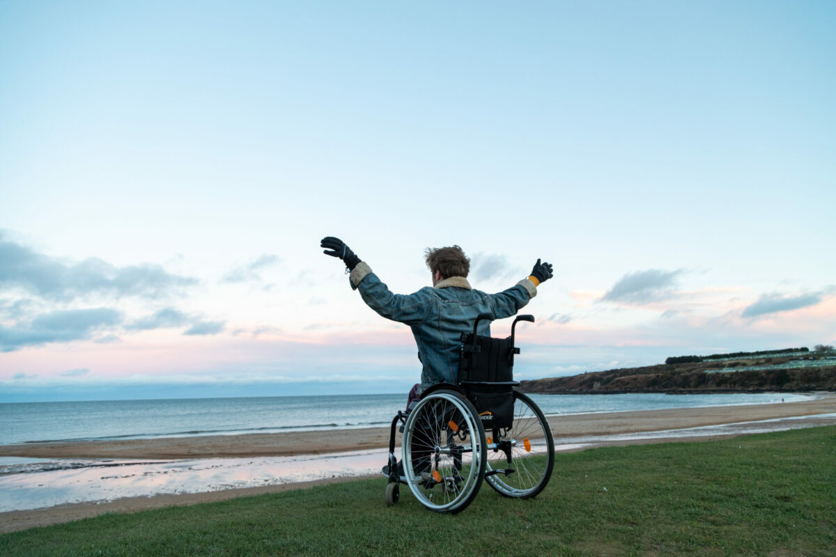A young person in a wheelchair with their arms above their head looking out over the beach.