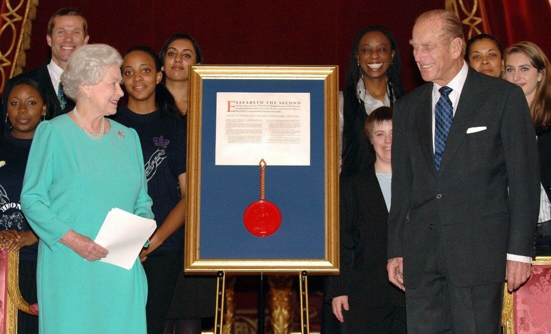 The Queen and Prince Phillip standing next to a frame with a document.