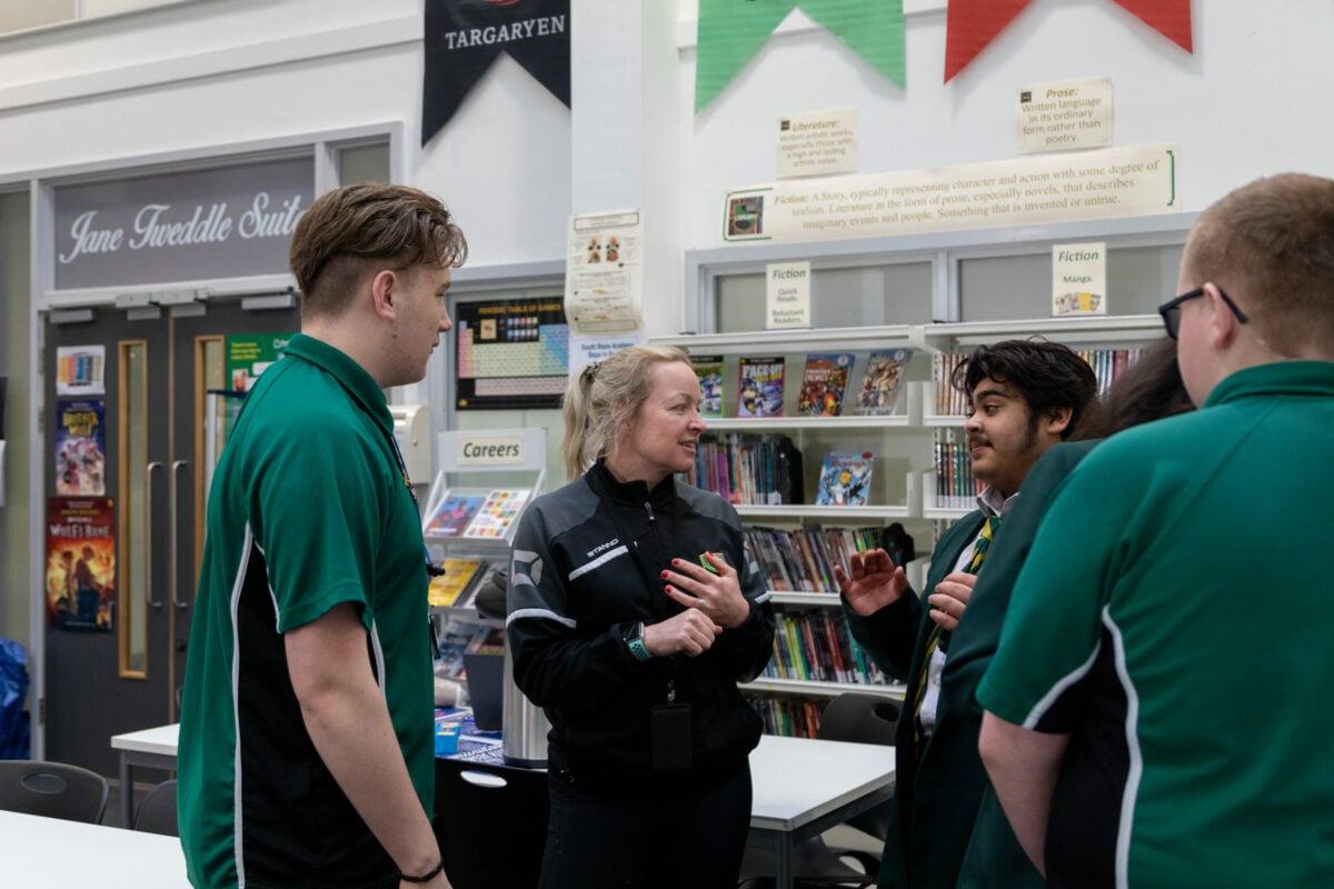 A staff member in a black sports-style jacket speaks with a small group of students wearing green uniforms inside a school building. They are gathered around a counter in an area with posters, noticeboards, and bookshelves, including signs for careers and a reading suite. The setting appears to be a school information or common area.