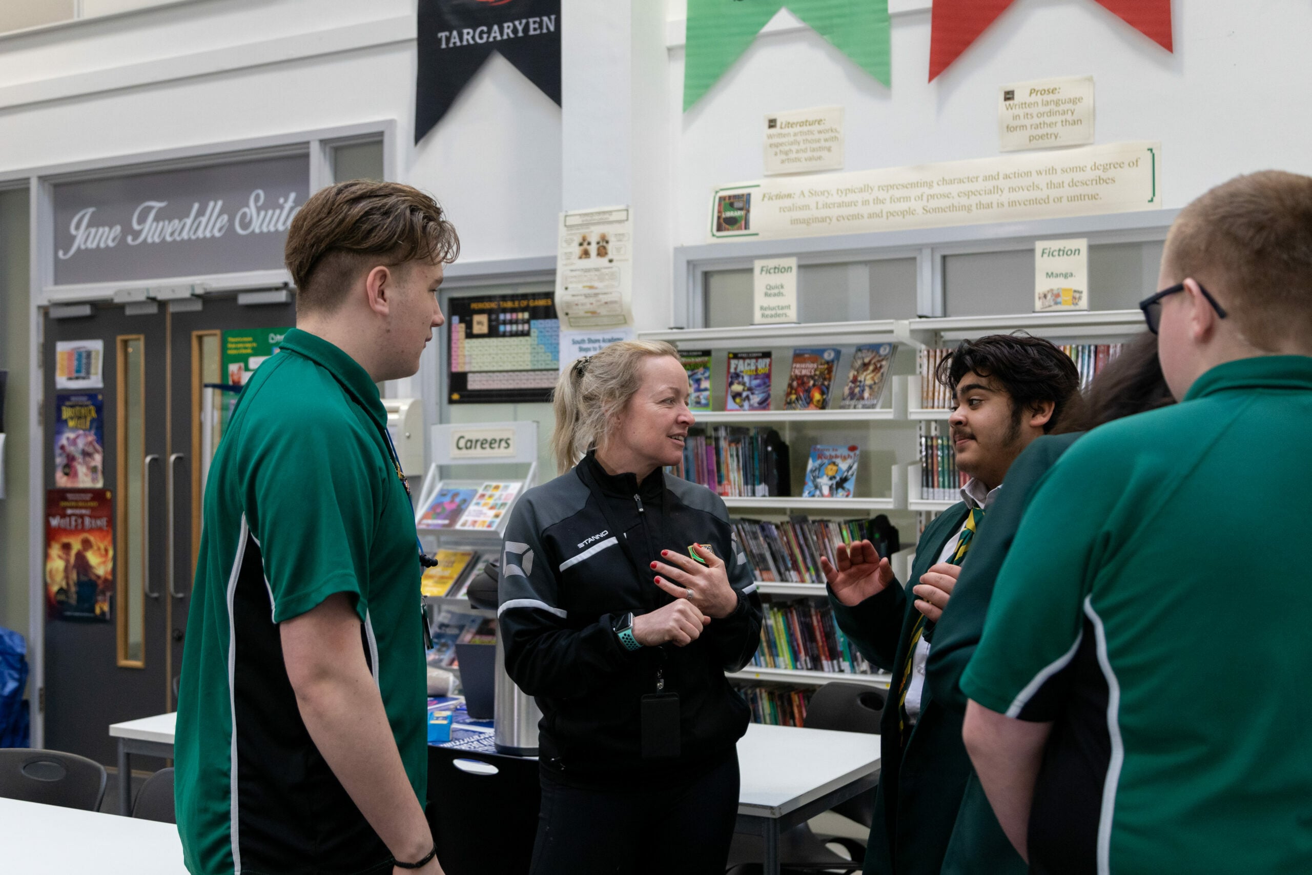 A staff member in a black jacket speaks with a small group of students wearing green uniforms inside a school building.