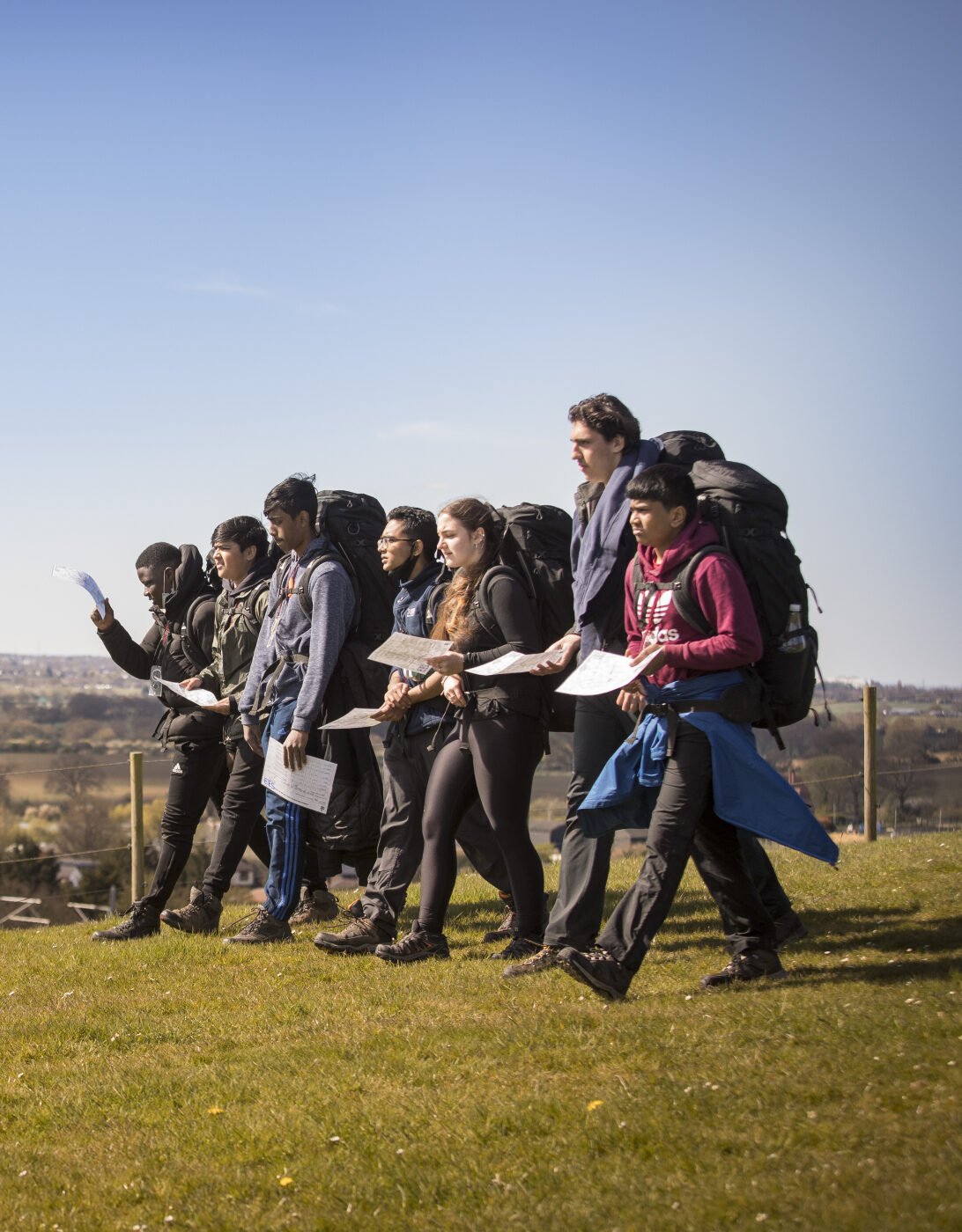A group of young people walk together across a grassy hilltop on a sunny day, holding maps and wearing outdoor clothing and backpacks. They appear to be taking part in an orienteering or hiking activity, with wide countryside views stretching out behind them under a clear blue sky.