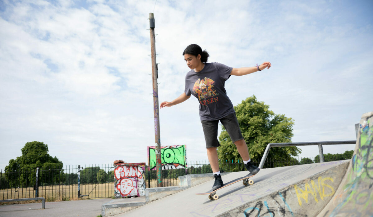 Josie skateboarding down a ramp at a graffiti-covered skate park, with trees and a partly cloudy sky in the background.