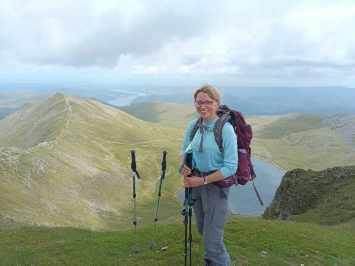 Hannah has blonde hair and pink glasses, she's carrying a rucksack and hiking poles and is standing on top of a mountain.