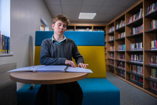 A young person sitting in a library reading a book in Braille.