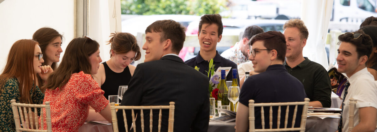 A group of people seated around a round table at an indoor event, engaged in conversation. The table is covered with a light-colored tablecloth and decorated with flowers and glassware. Wooden chairs with light backs surround the table, and the background shows bright natural light coming through large windows.