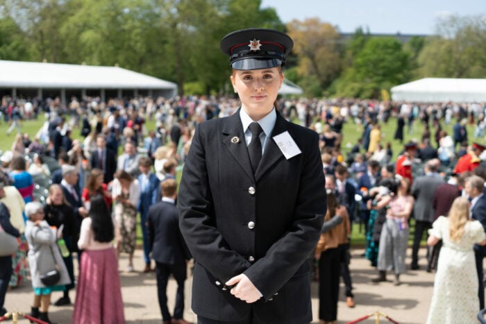 Ffion standing on the steps of Buckingham Palace gardens in front of a crowd of young people and their parents. She is wearing her fire service uniform and looking directly at the camera.