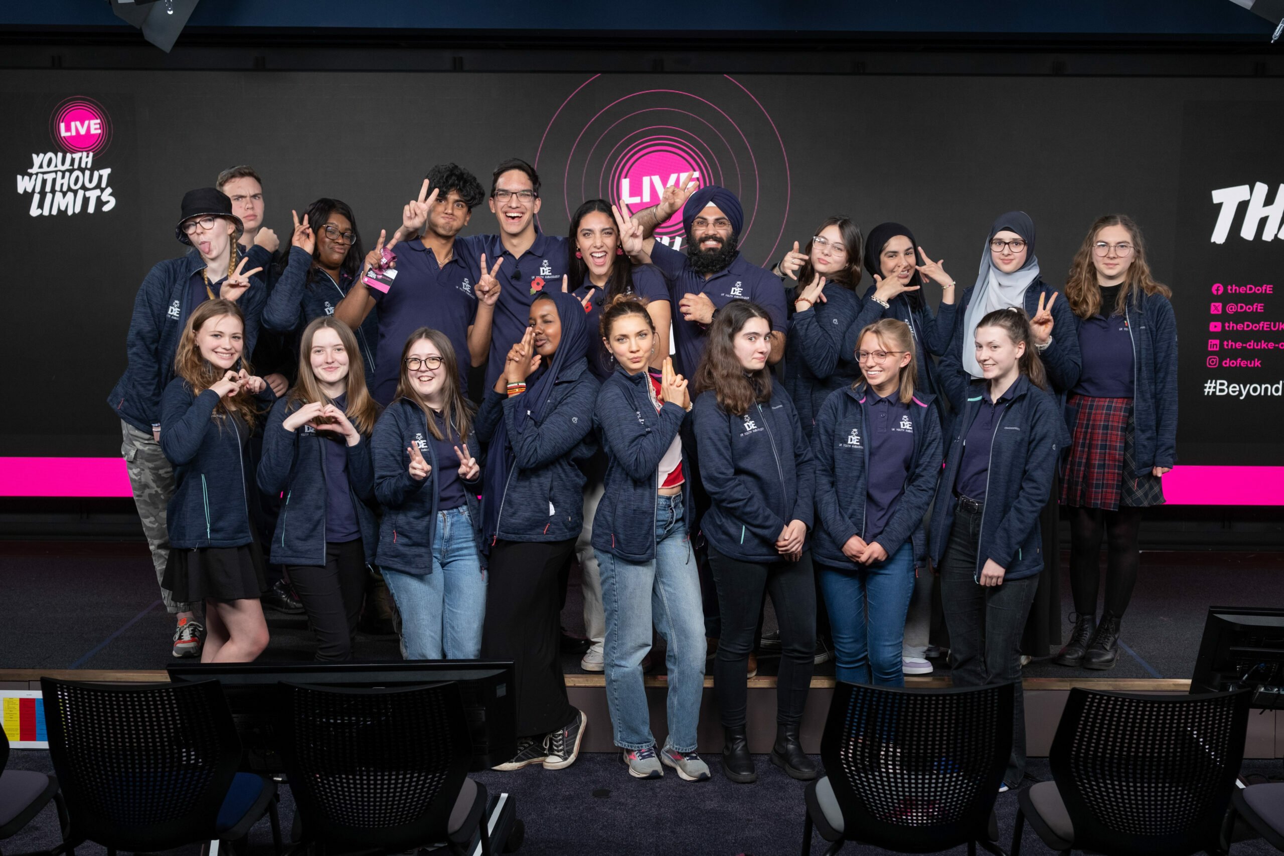 A group of 19 DofE UK Youth Ambassadors standing on stage at the DofE's Youth Without Limits LIVE event. They are all wearing matching navy fleeces, doing different poses and looking directly to the camera.