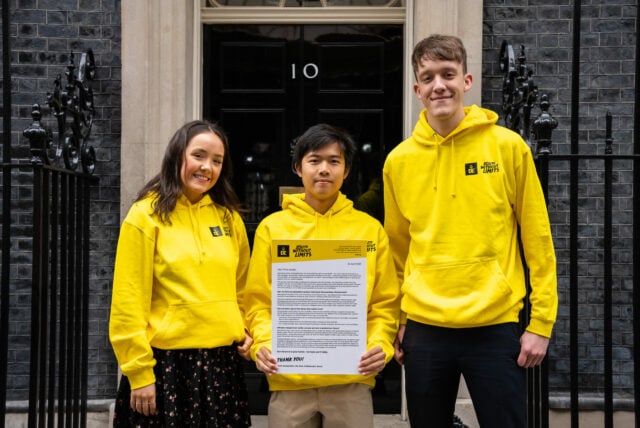 Three young people all wearing yellow hoodies standing outside the black10 Downing Street door. They are holding a large letter addressed to the Prime Minister.