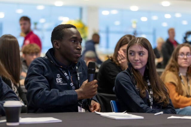 A young Youth Ambassador sitting at a conference table and talking into a microphone. He is sitting next to another young person who is looking directly at him.