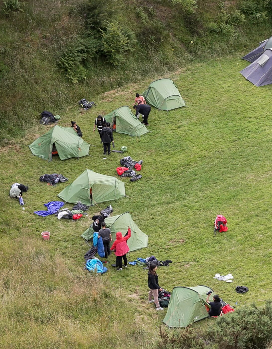 An aerial shot of a campsite. The tents are on grass, in a circle with a large space in the middle. There are young people next to their tents setting them up.