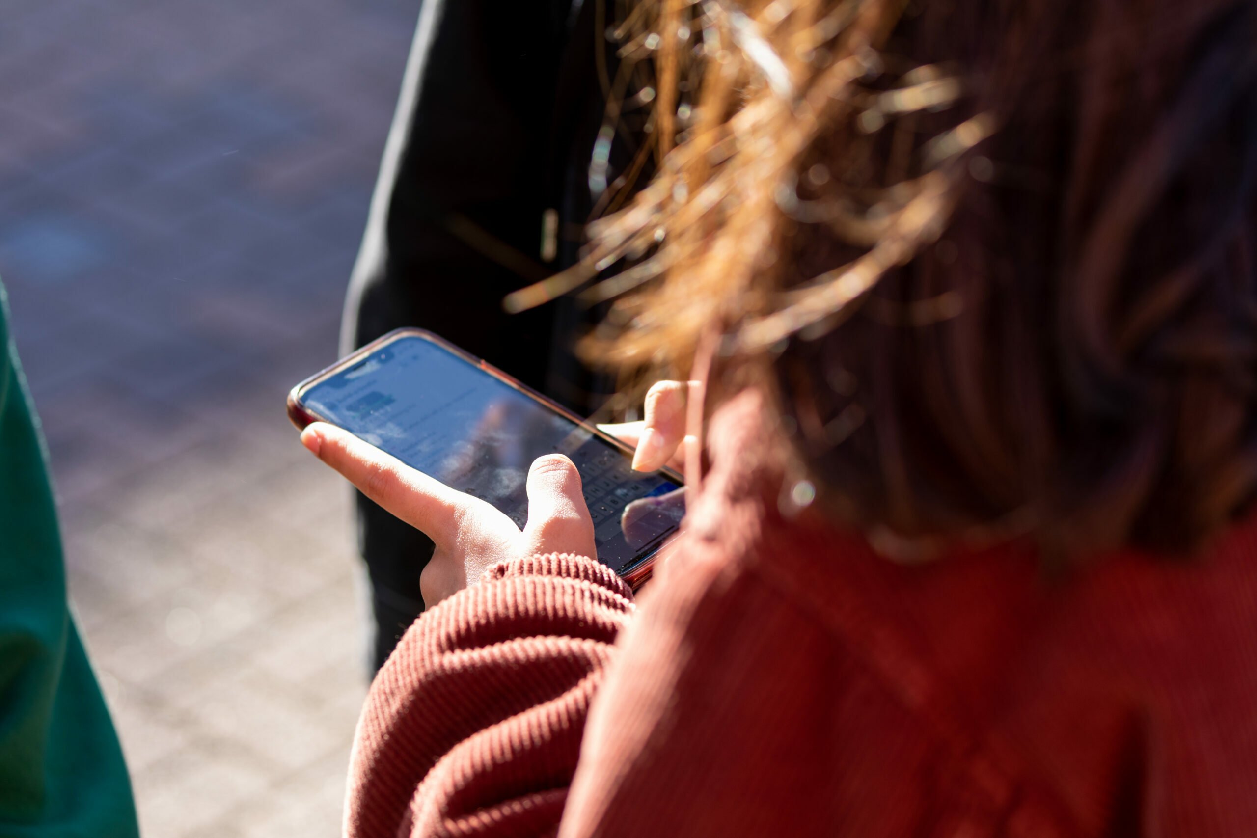 A young person holding a mobile phone. The photo is taken over her shoulder and we can just see her hands and the mobile.