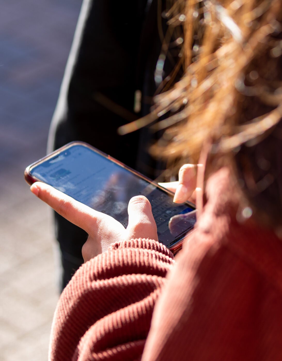 A young person holding a mobile phone. The photo is taken over her shoulder and we can just see her hands and the mobile.