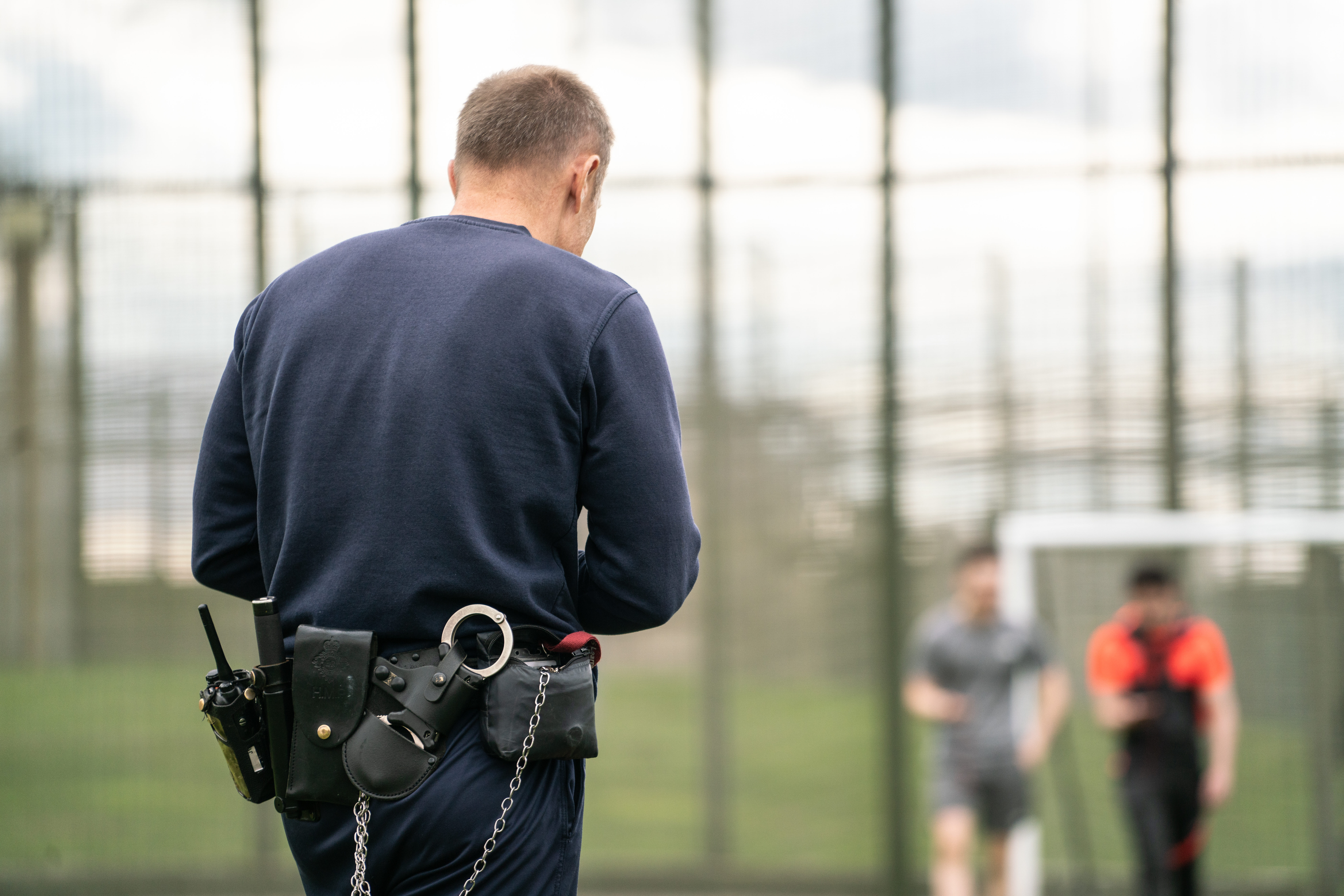 A prison officer supervising some young offenders while they play football. He has his back to the camera and we can see prison handcuffs on his belt.