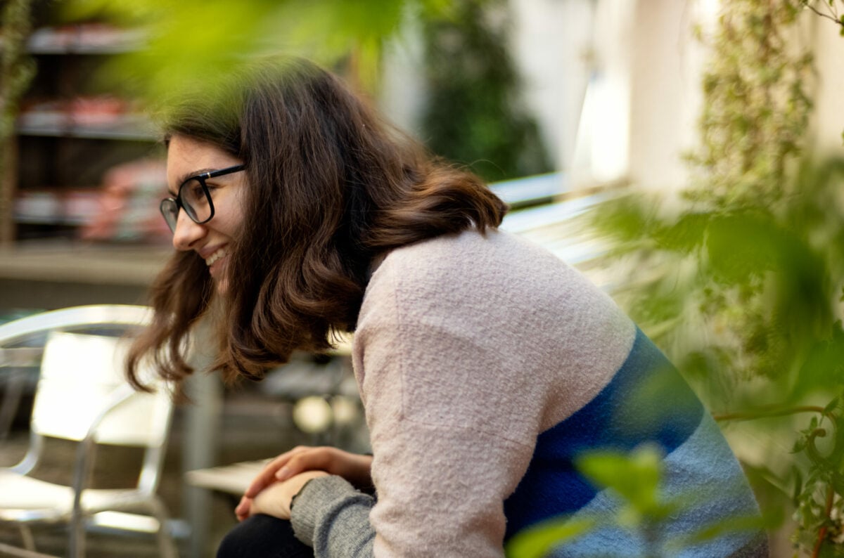 A young person wearing glasses laughing away from the camera. She is facing side on and is outside, she is partially blocked by leaves.