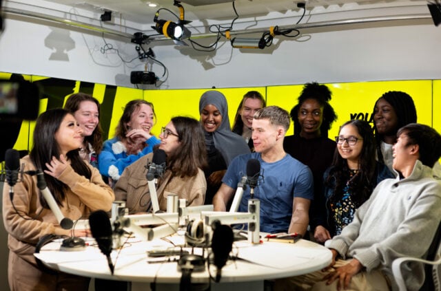 11 young people all sitting and standing around a table with podcast microphones on it. They are laughing and looking at each other. The backdrop is a yellow banner that reads 