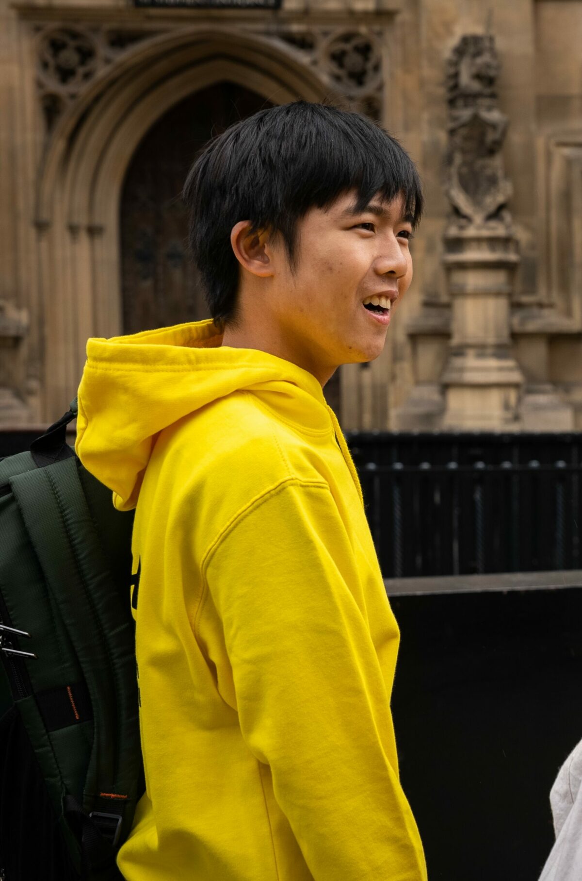 A young person wearing a yellow hoodie laughing away from the camera. He is standing outside Westminster Abbey and looking at someone not in shot.
