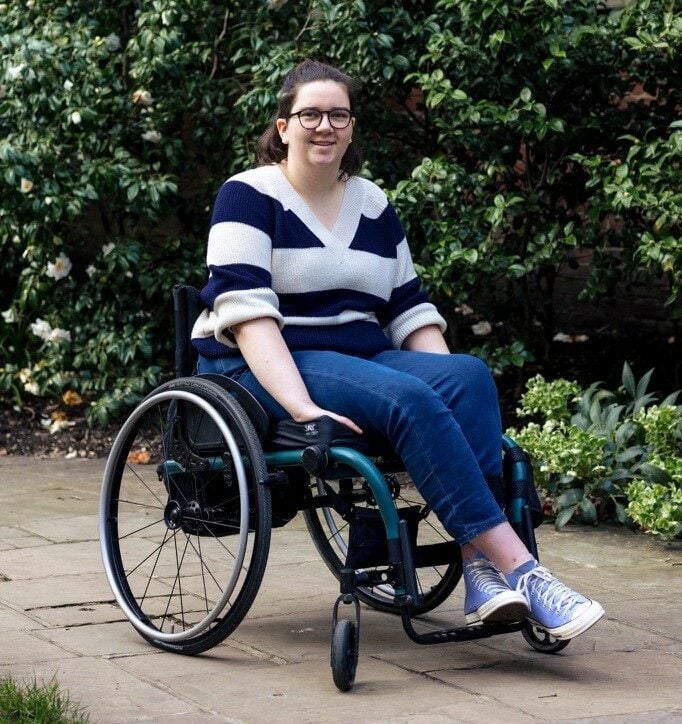 Alice has short brown hair, uses a wheelchair and is wearing a black and white striped jumper.