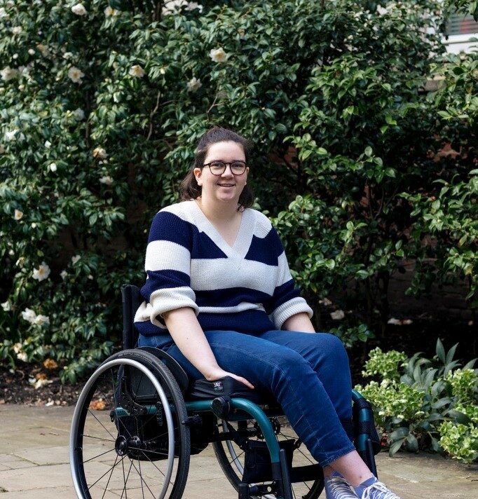 A young person smiling directly into the camera. They are outside and sitting in front of green bushes.