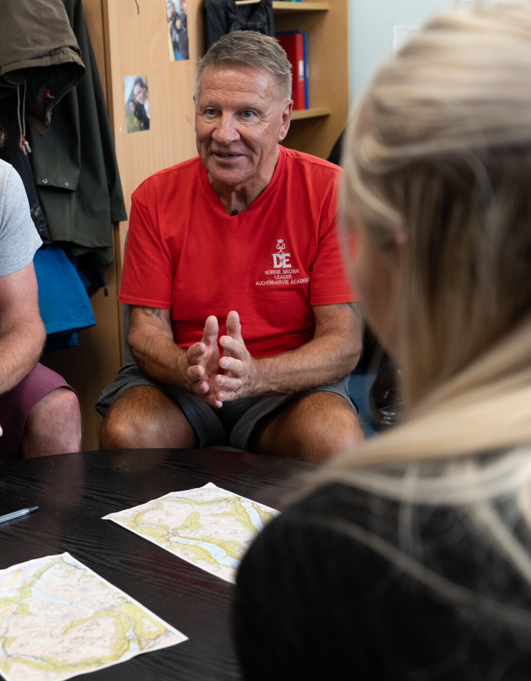 Image of 2 men sitting around a table with sheets of paper in front of them. They are talking to 2 young people and we can see the back of their heads.
